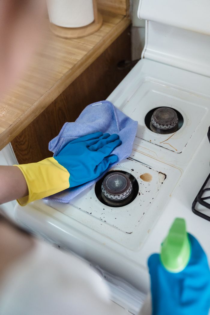 about-01 A person wearing rubber gloves cleaning a dirty gas stove with a cloth and spray bottle.